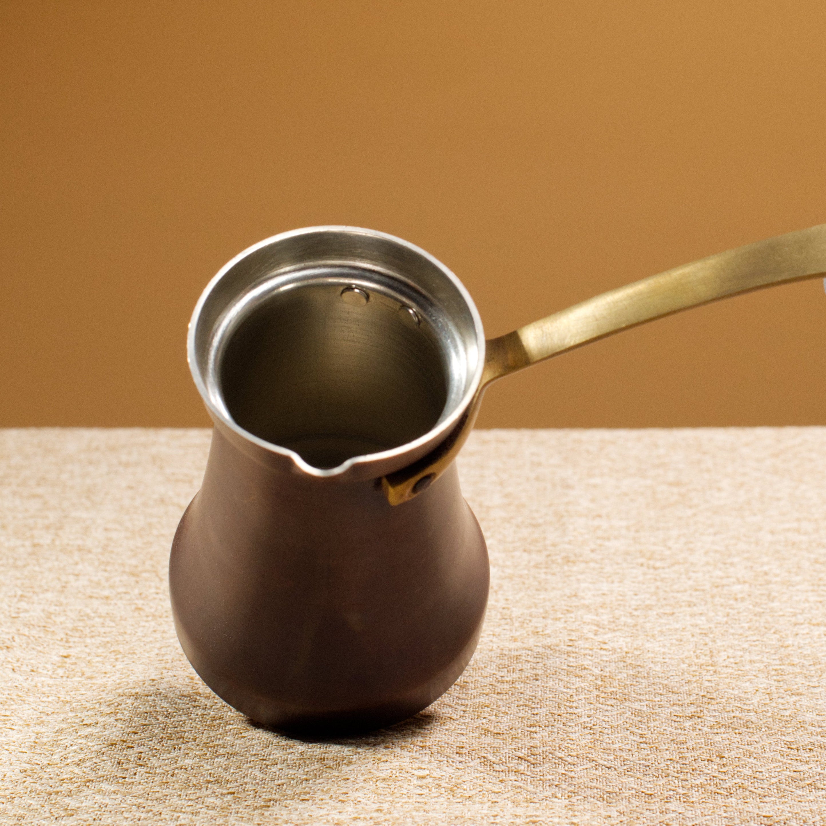Brown coffee pot with brass handle on a beige surface against a yellow background